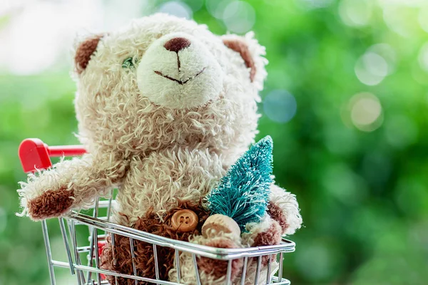 Smiling teddy bear doll sitting in mini shopping cart or trolley with Christmas tree against blurred natural green background for children gift