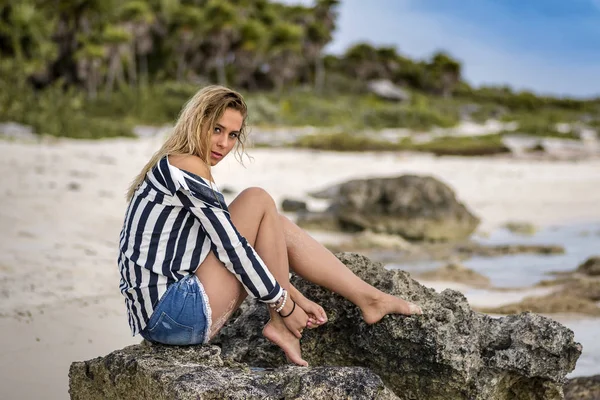Güzel genç sarışın Avrupa kız kireç taş kayanın üzerinde ıslak saçlı, çizgili gömlek bir Karayip plajda kameraya bakarak oturuyor