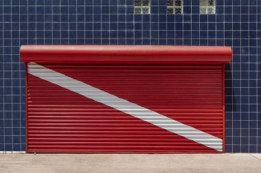 Closed store shutter painted to red scuba diving flag with a wall covered with blue tiles clipart