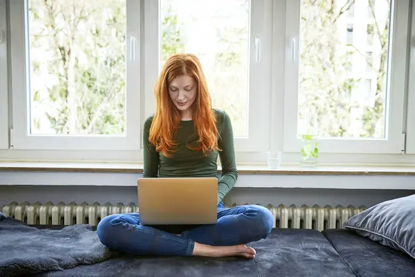 红发女人坐在窗前使用笔记本电脑 — 图库照片