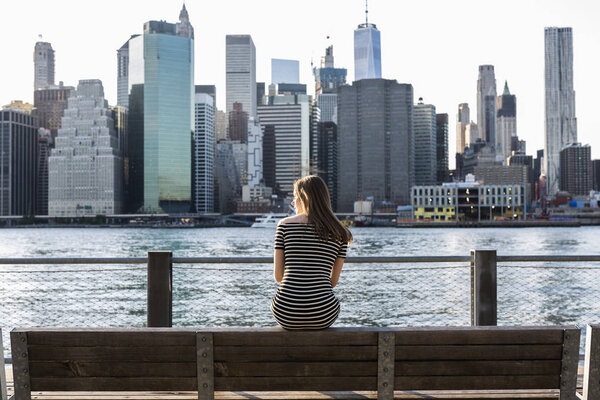 USA, New York, Brooklyn, back view of woman sitting on bench in front of East River and skyline of Manhattan