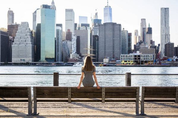 USA, New York, Brooklyn, back view of woman sitting on bench in front of East River and skyline of Manhattan