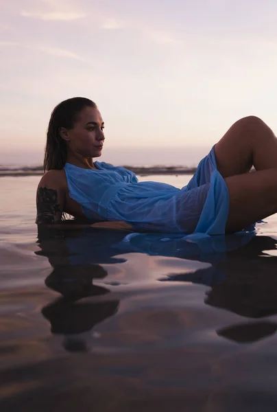日落前 身穿蓝色连衣裙 纹身的年轻女子躺在海滨的水里 — 图库照片