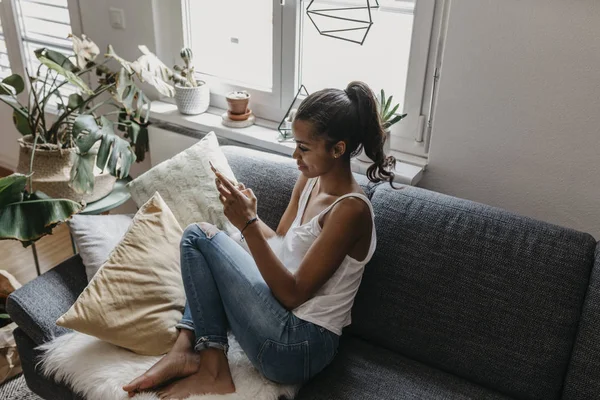 Молода жінка сидить на дивані вдома за допомогою мобільного телефону — стокове фото