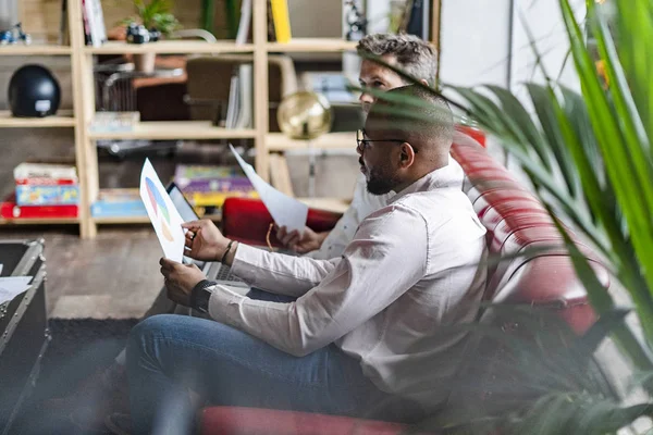 Zwei Geschäftsleute nutzen Laptop und diskutieren Dokumente auf Sofa im Loft-Büro — Stockfoto