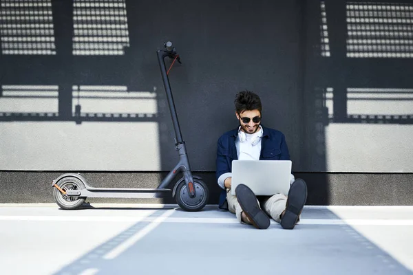 Hombre joven relajado sentado en el suelo, además de su scooter eléctrico utilizando el ordenador portátil - foto de stock