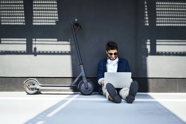 Rilassato giovane seduto a terra oltre al suo scooter elettrico utilizzando il computer portatile — Foto stock