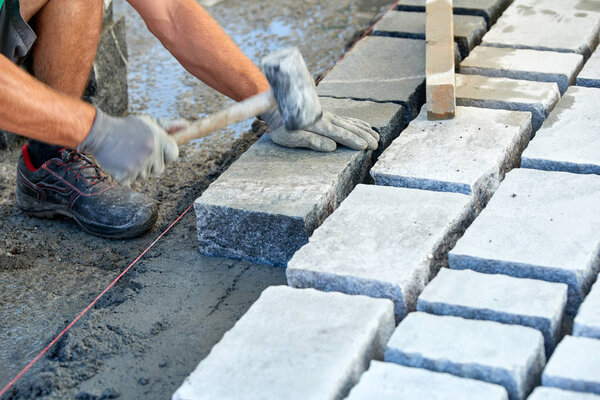 A workman's gloved hands use a hammer to place stone pavers. Worker creating pavement using cobblestone blocks and granite stones.