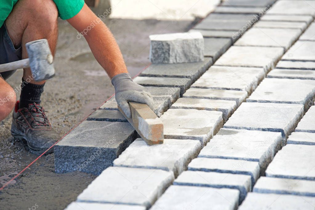 A workman's gloved hands use a hammer to place stone pavers. Worker creating pavement using cobblestone blocks and granite stones.