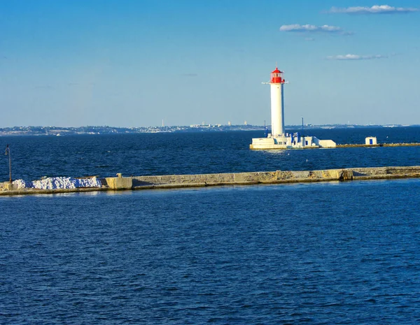 A lighthouse in the sea at the entrance to the port against the backdrop of a beautiful evening blue sky in soft colors. Summer seascape. White lighthouse with a red top. Black Sea. Seaport of Odessa