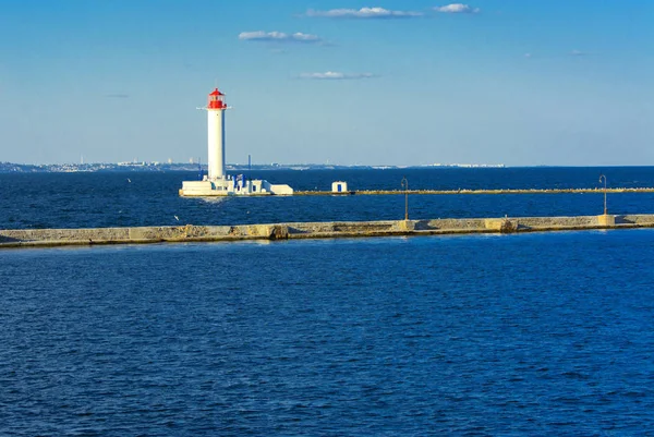 A lighthouse in the sea at the entrance to the port against the backdrop of a beautiful evening blue sky in soft colors. Summer seascape. White lighthouse with a red top. Black Sea. Seaport of Odessa