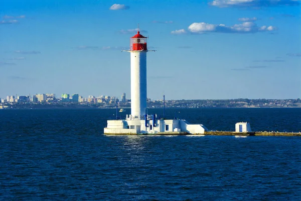 A lighthouse in the sea at the entrance to the port against the backdrop of a beautiful evening blue sky in soft colors. Summer seascape. White lighthouse with a red top. Black Sea. Seaport of Odessa