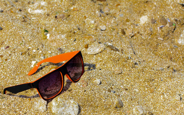 Sunglasses are lost and lie on the beach by the sea. Black with orange edging glasses on the background of yellow sand on the shore. Sunglasses are reflected in the golden wet sand, as in a mirror