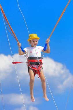 Küçük bir kız yüksek lastik iplere mavi gökyüzü ve beyaz bulutlar ile bir trambolin üzerinde atlar. Macera ve doğa sporları. Yaz tatili, atlama kavramı.
