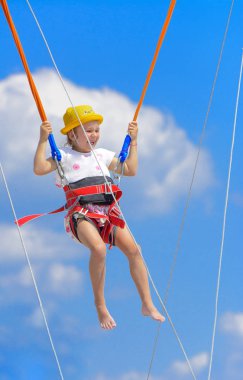 Küçük bir kız yüksek lastik iplere mavi gökyüzü ve beyaz bulutlar ile bir trambolin üzerinde atlar. Macera ve doğa sporları. Yaz tatili, atlama kavramı.