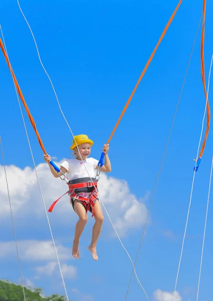 A little girl jumps high on a trampoline with rubber ropes against the blue sky and white clouds. Adventures and extreme sports. Concept of summer vacation, jumping.