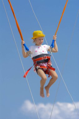 Küçük bir kız yüksek lastik iplere mavi gökyüzü ve beyaz bulutlar ile bir trambolin üzerinde atlar. Macera ve doğa sporları. Yaz tatili, atlama kavramı.