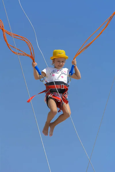 A little girl jumps high on a trampoline with rubber ropes against the blue sky and white clouds. Adventures and extreme sports. Concept of summer vacation, jumping.