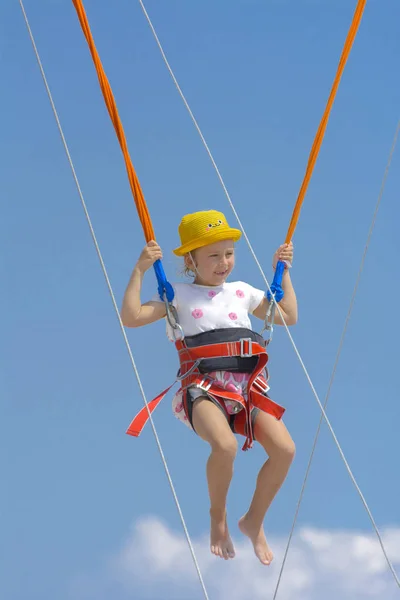 A little girl jumps high on a trampoline with rubber ropes against the blue sky and white clouds. Adventures and extreme sports. Concept of summer vacation, jumping.