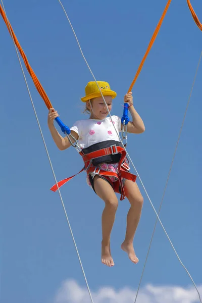 A little girl jumps high on a trampoline with rubber ropes against the blue sky and white clouds. Adventures and extreme sports. Concept of summer vacation, jumping.