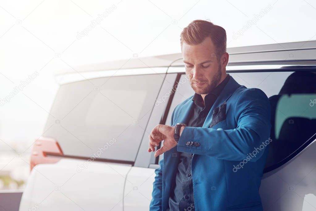Modern etrepreneur. Waist up of a handsome self assertive businessman checking the time and leaning on his car while standing in the parking lot