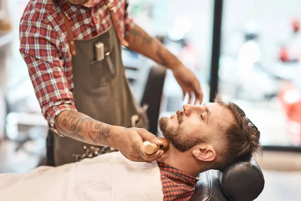 Professional salon. Masterful barber finishing shaving the beard of a handsome young bearded man sittiing in the chair