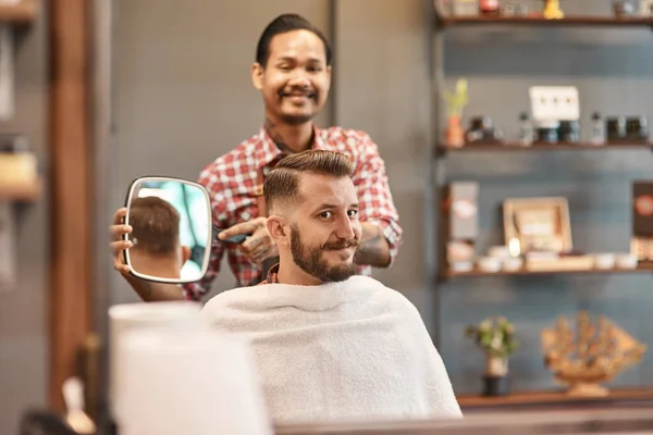 Lokks great. Cheerful bearded man sitting in the barbershop and looking in the mirror while a smiling barber standing in the background