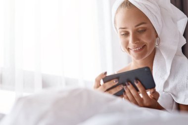 Duygu dolu. Yatakta dinleniyor ve mutlu hissederken onun smartphone kullanarak neşeli genç güzel kadın