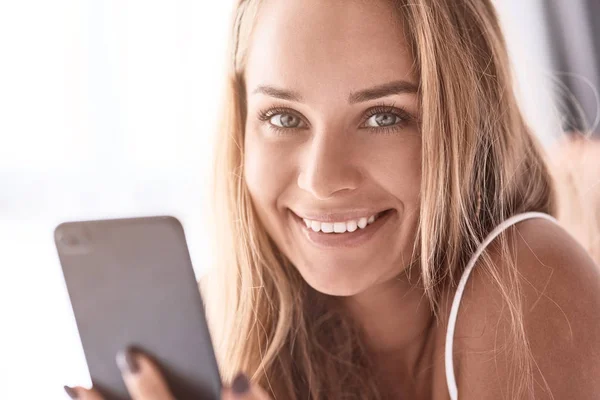 Share your emotions. Joyful beautiful young woman smiling and looking at you while holding her smartphone