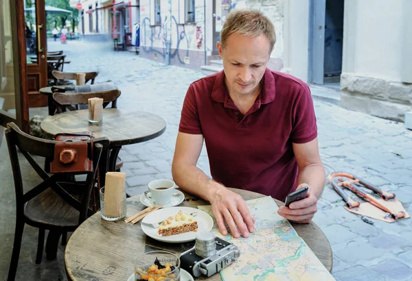 The man is holding a smartphone. He is sitting at the cafe table on the street. On the table there is a vintage camera, a tourist map, a coffee and a piece of cake on a plate.