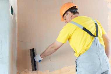 İşçi macun duvardaki bir fiberglas ağ üzerinde uyguluyor. O geniş bir spatula kullanıyor.