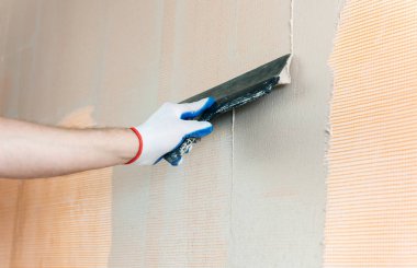 Bir işçi macun duvardaki bir fiberglas ağ üzerinde uyguluyor. O geniş bir spatula kullanıyor.