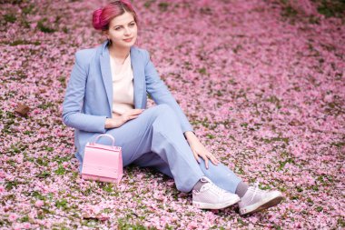 arka plan bahçe pembe yaprakları üzerinde oturan mavi şık elbiseli pembe saçlı kız