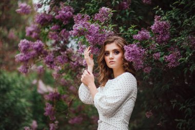 Baharda leylak çalısı yanında beyaz bir gece elbisesi giymiş bir kızın portresi.