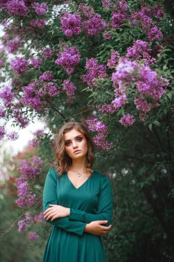 Baharda yeşil bir elbiseyle çiçek açmış leylak çalılarının arkasında poz veren güzel bir kızın şık portresi.