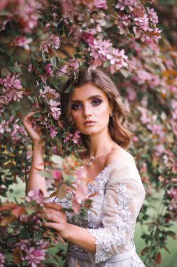 Beyaz narin elbiseli güzel romantik bir kız baharda pembe çiçeklerden oluşan bir çalılığın yanında duruyor ve çiçeklerle dolu bir dala dokunuyor.