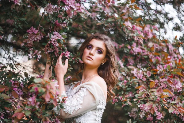 在一丛丛粉红色的花旁边 站着一位穿着白色精致衣服的美丽浪漫的姑娘 她摸着枝头上的花 — 图库照片
