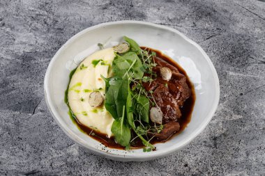 Patates ve lahana soslu dana yanakları, beyaz bir tabakta otlarla süslenmiş.