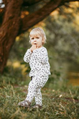 Doğanın arka planına karşı güzel beyaz bir takım elbiseyle yürüyen tatlı bir çocuk. Bir yaşında bir çocuk öğreniyor ve dışarıda oynuyor..