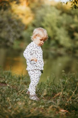 Doğanın arka planına karşı güzel beyaz bir takım elbiseyle yürüyen tatlı bir çocuk. Bir yaşında bir çocuk öğreniyor ve dışarıda oynuyor..