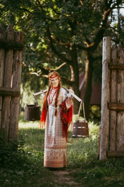 Uzun sarı saçlı, kahverengi gözlü, beyaz ve kırmızı işlemeli takım elbiseli ve omuzlarında boyundurukla güzel bir Slav kız. Ukrayna bölgesinin geleneksel kıyafetleri. Yaz günü.