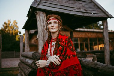 Beyaz ve kırmızı işlemeli takım elbiseli, uzun sarı saçlı ve kahverengi gözlü güzel bir Slav kız tahta bir evin yanında oturuyor. Ukrayna bölgesinin geleneksel kıyafetleri. Yaz günü.