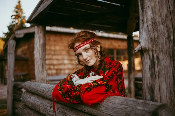 一位漂亮的斯拉夫姑娘坐在一座木头房子旁边 她的头发是金色的 眼睛是棕色的 身穿白色和红色绣花的西服 — 图库照片