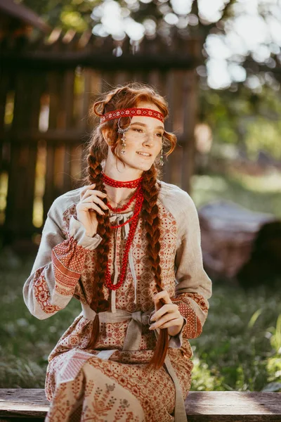 白と赤の刺繍のスーツで長いブロンドの髪と茶色の目を持つ美しいスラブの少女は 木製のフェンスの横に座っています ウクライナの地域の伝統的な服 夏の日 — ストック写真