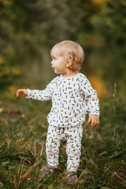 Doğanın arka planına karşı güzel beyaz bir takım elbiseyle yürüyen tatlı bir çocuk. Bir yaşında bir çocuk öğreniyor ve dışarıda oynuyor..