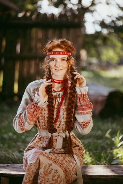 白と赤の刺繍のスーツで長いブロンドの髪と茶色の目を持つ美しいスラブの少女は 木製のフェンスの横に座っています ウクライナの地域の伝統的な服 夏の日 — ストック写真