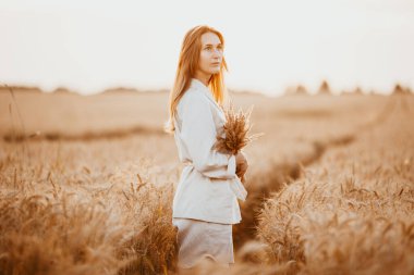 Uzun kıvırcık saçlı ve açık beyaz takım elbiseli genç bir kız buğday tarlasında kameraya poz veriyor, günbatımının arka planında.