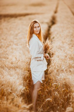 Uzun kıvırcık saçlı ve açık beyaz takım elbiseli genç bir kız buğday tarlasında kameraya poz veriyor, günbatımının arka planında.