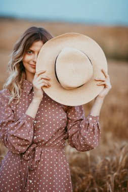 Beyaz benekli kahverengi elbiseli genç bir kadın elinde şapkayla buğday tarlasında duruyor.