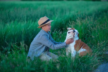 Onun İngilizce boğa köpek yeşil çavdar çayır üzerinde küçük çocuk oturma dururken. Şık giysiler ve rustik retro şapka giyen en iyi arkadaşıyla poz Handsom çocuk.